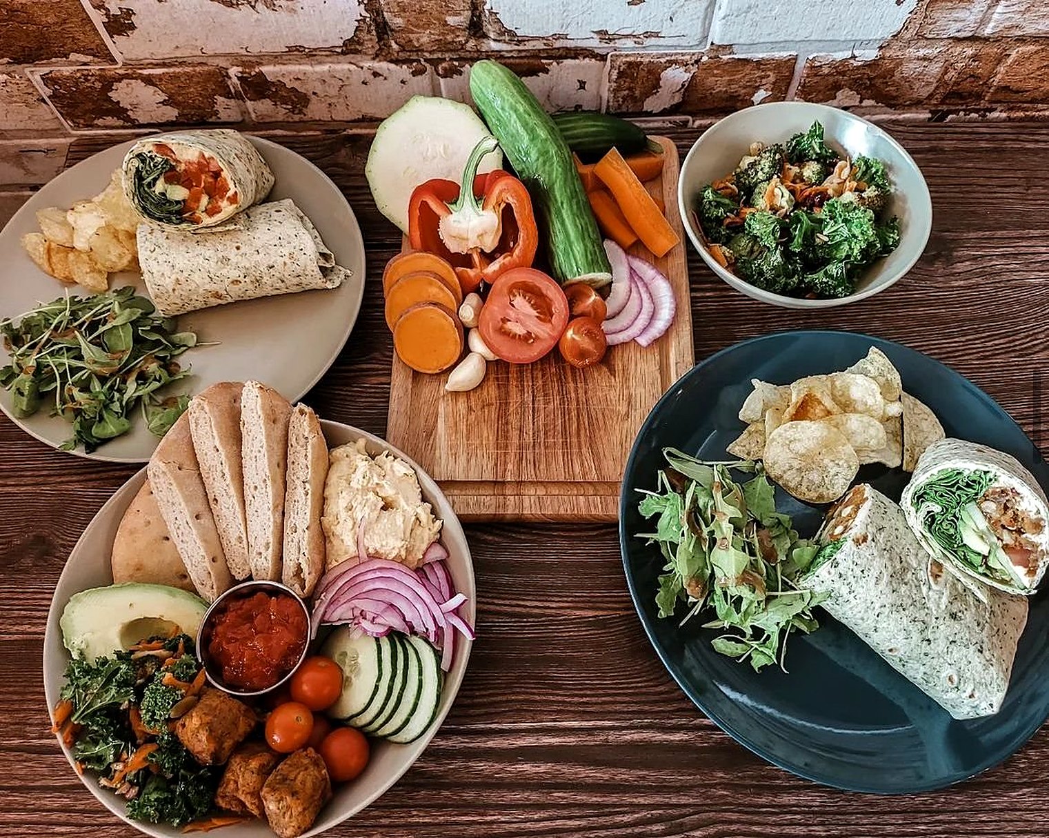 A table of food seen from above: wraps, flatbread with hummus, salad leaves, avocado, cherry tomatoes and a board of chopped vegetables