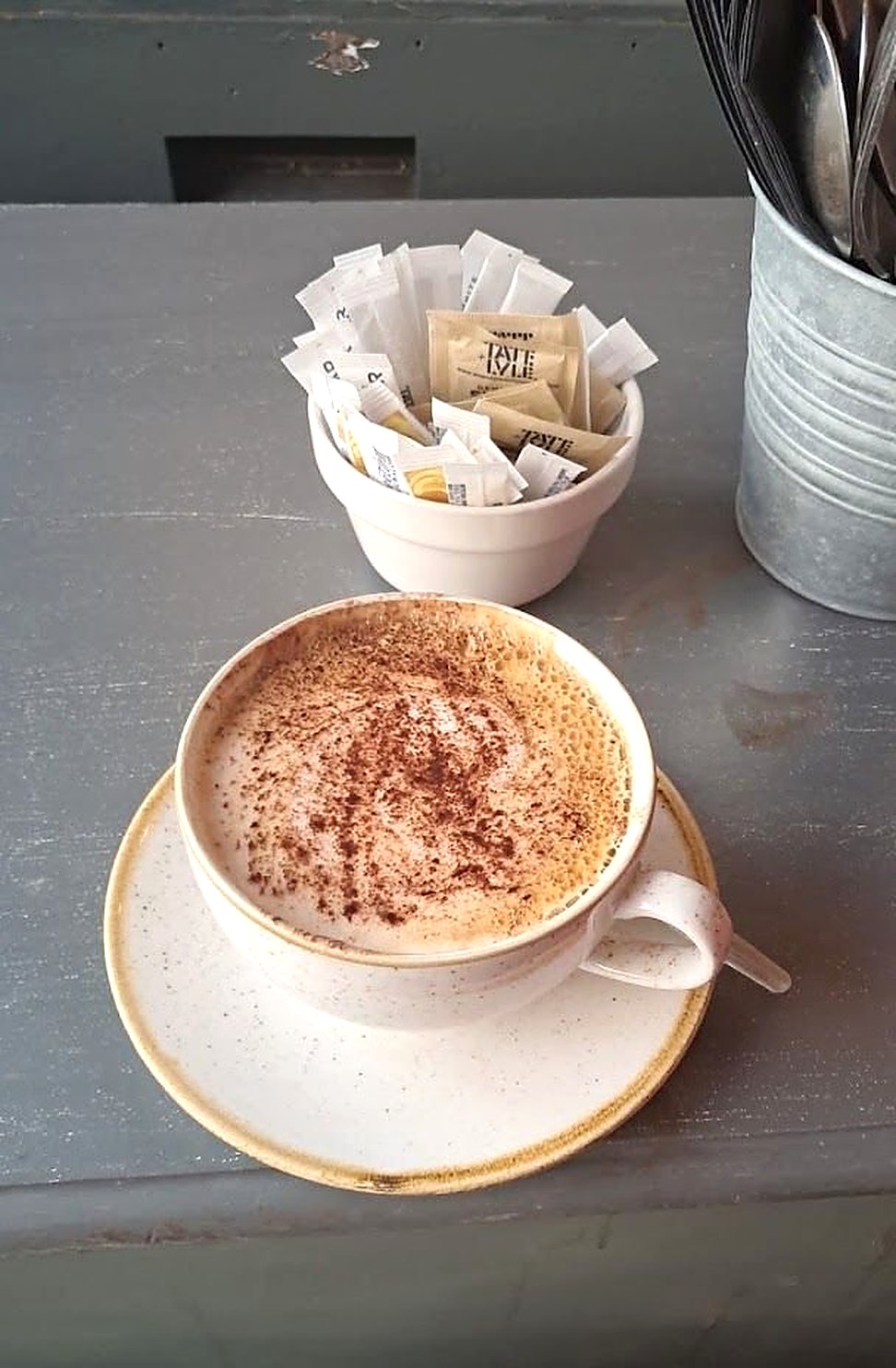 A cappuccino dusted with chocolate in a cream cup and saucer, beside a pot of sugar sachets