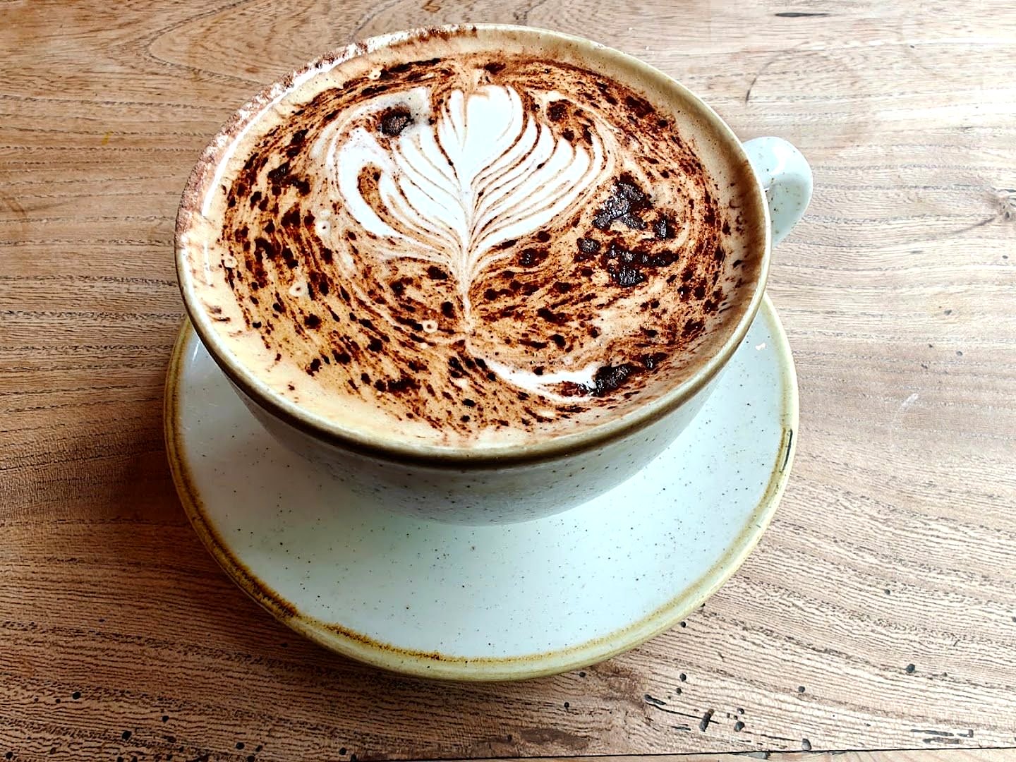 A latte with a rosetta of milk foam, dusted with chocolate, in a speckled cup on a wooden table